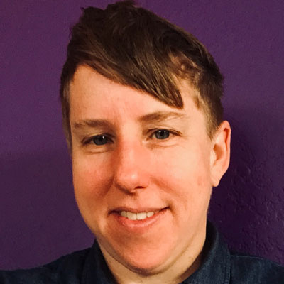 A photo of gs practitioner and teacher Amanda Ream. A white person with short hair, and bangs that swoop to the right of the photo is smiling into the camera. They are wearing a dark blue shirt and are in front of a wall that has been painted a deep shade of purple.