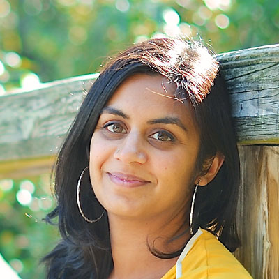 A photo of gs teacher and practitioner Fayza Bundalli. A brown woman with pink lipstick is leaning against a wooden railing with trees behind her. She is wearing pink lipstick, silver hoop earrings, and a yellow and white top.