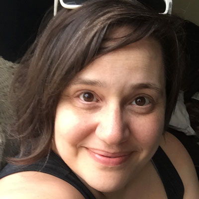 A photo of gs teacher and practitioner Briana Herman-Brand. A white woman is turning her head to the right to smile into the camera directly. She has brown hair in an asymmetrical haircut and you can see the rims of white glasses on top of her head, and is wearing a black sleeveless shirt.
