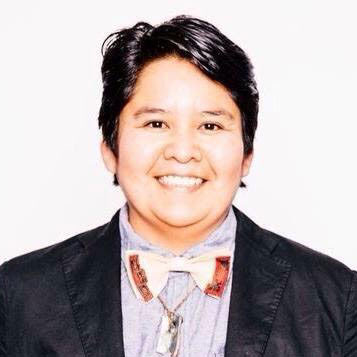 A picture of gs teacher and practitioner Nazbah Tom. A light brown person with their short black hair parted is smiling into the camera, wearing a blue button up shirt, a black jacket, and a white and red bowtie with a silver necklace underneath that.