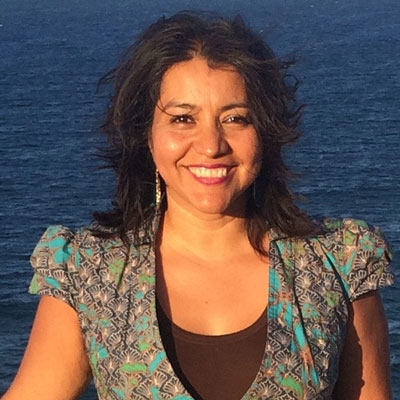 A photo of gs teacher and practitioner Donaji Lona Esteva. A brown woman is smiling into the camera with the ocean behind her. She is wearing a multicololored blue, orange, and black dress, red lipstick, and long gold earrings.