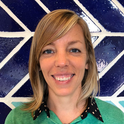 A photo of gs teacher and practitioner Hilary Moore. A white woman with medium-length blond hair smiles into the camera. She is wearing a green shirt with a black collar that has green polka dots. Behind her is a wall made of blue tiles and white grout.