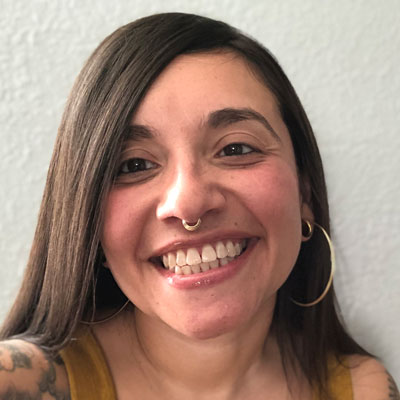 A picture of gs teacher and practitioner Natalíe Lorraine Ortega. A Latinx woman with long brown hair, a gold septum ring, and hoop earrings smiles into the camera. Behind her is a white wall.