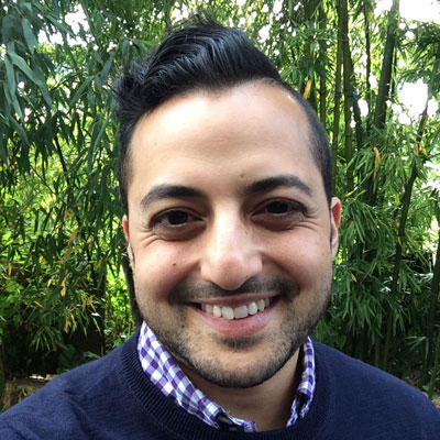 A photo of gs teacher and practitioner Nathan Shara. A Brown man is smiling into the camera with short black hair slicked back and to the side and a small beard, wearing a blue sweater over a blue plaid collared shirt. Behind him is a grove of bamboo stalks.