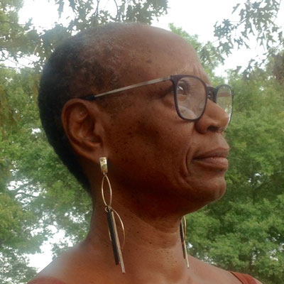 A photo of gs practitioner and teacher Alta Starr. A Black woman with short salt and pepper black hair is looking ahead of her, towards the right of the photo. She is wearing black-rimmed glasses, and long gold dangly earrings. Behind her is the sky and trees full of green leaves.