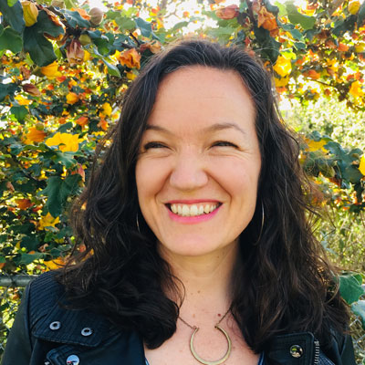 A photo of gs teacher and practitioner Lara Barth. A white person with long wavy black hair smiles and looks to the right of the photo. She is wearing gold hoop earrings and a gold necklace and a black jacket. Behind her are trees and a fence with yellow flowers.