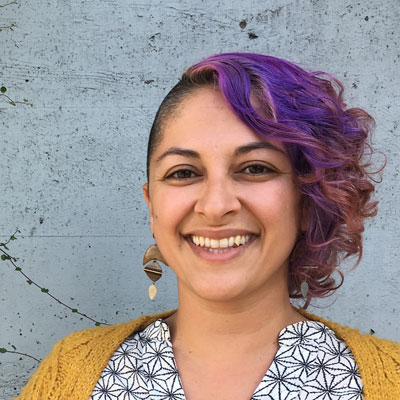 A photo of gs Program Associate, teacher, and practitioner Mahfam Malek. A light brown-skinned person is looking at and smiling into the camera. They have short hair on the sides and long curly purple and pink hair on the top. They are weaaring dangly earrings consisting of a crescents and diamond shaped metals and rocks, a black and white blouse and a mustard yellow cardigan. They are standing infront of a light grey wall.