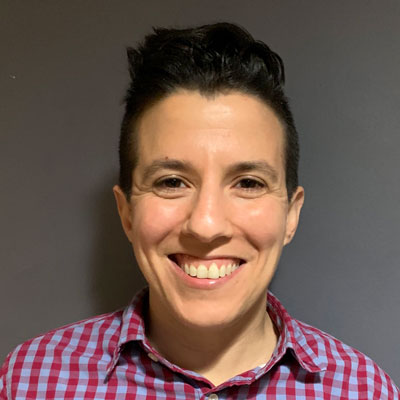 A photo of gs Resource Development Director and teacher, Danielle Feris. A white person with short black hair that is slightly longer on top is smiling infront of the camera. They are wearing a blue and red plaid button up shirt and are standing infront of a grey background.