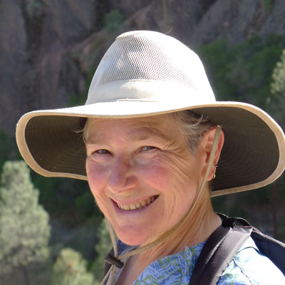 A photo of gs practitioner Elizabeth Ross. A white person is smiling into the camera, wearing a wide-brimmed light brown hat and a black backpack, and a blue shirt with a pattern on it.